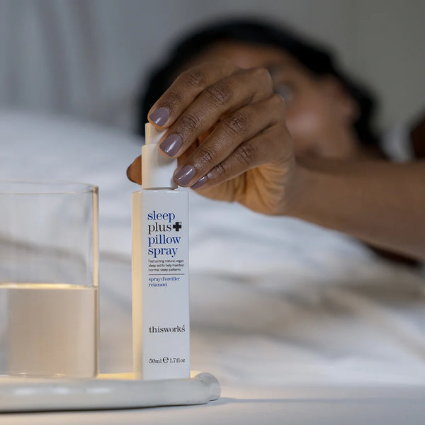 Persoon in bed plaatst This Works Sleep Plus+ Pillow Spray op nachtkastje