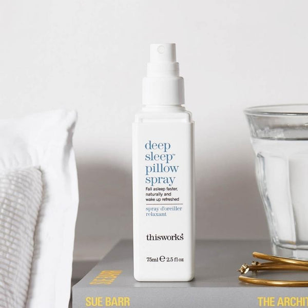 This Works Deep Sleep Pillow Spray 75 ml op een boek op een nachtkastje met een glas water en kussen op de achtergrond