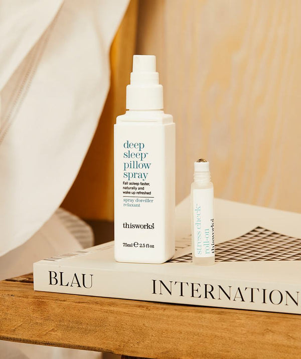 Sfeervol beeld van de This Works Deep Sleep Pillow Talk op een boek op een nachtkastje naast het bed, met een stuk van het kussen zichtbaar.